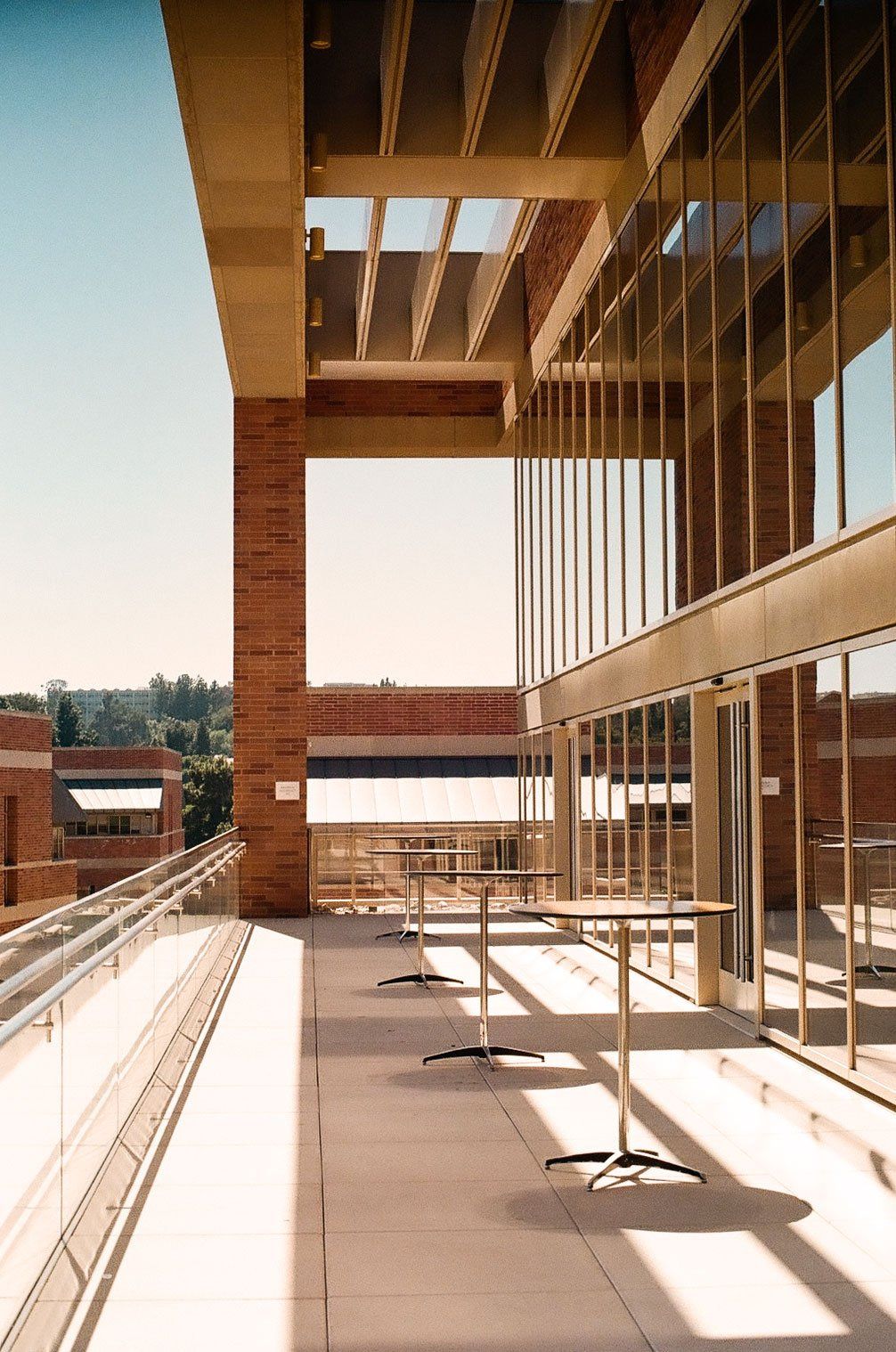 A quiet terrace in late afternoon sun: tall brick columns, a glass wall, and a few high tables casting long shadows.