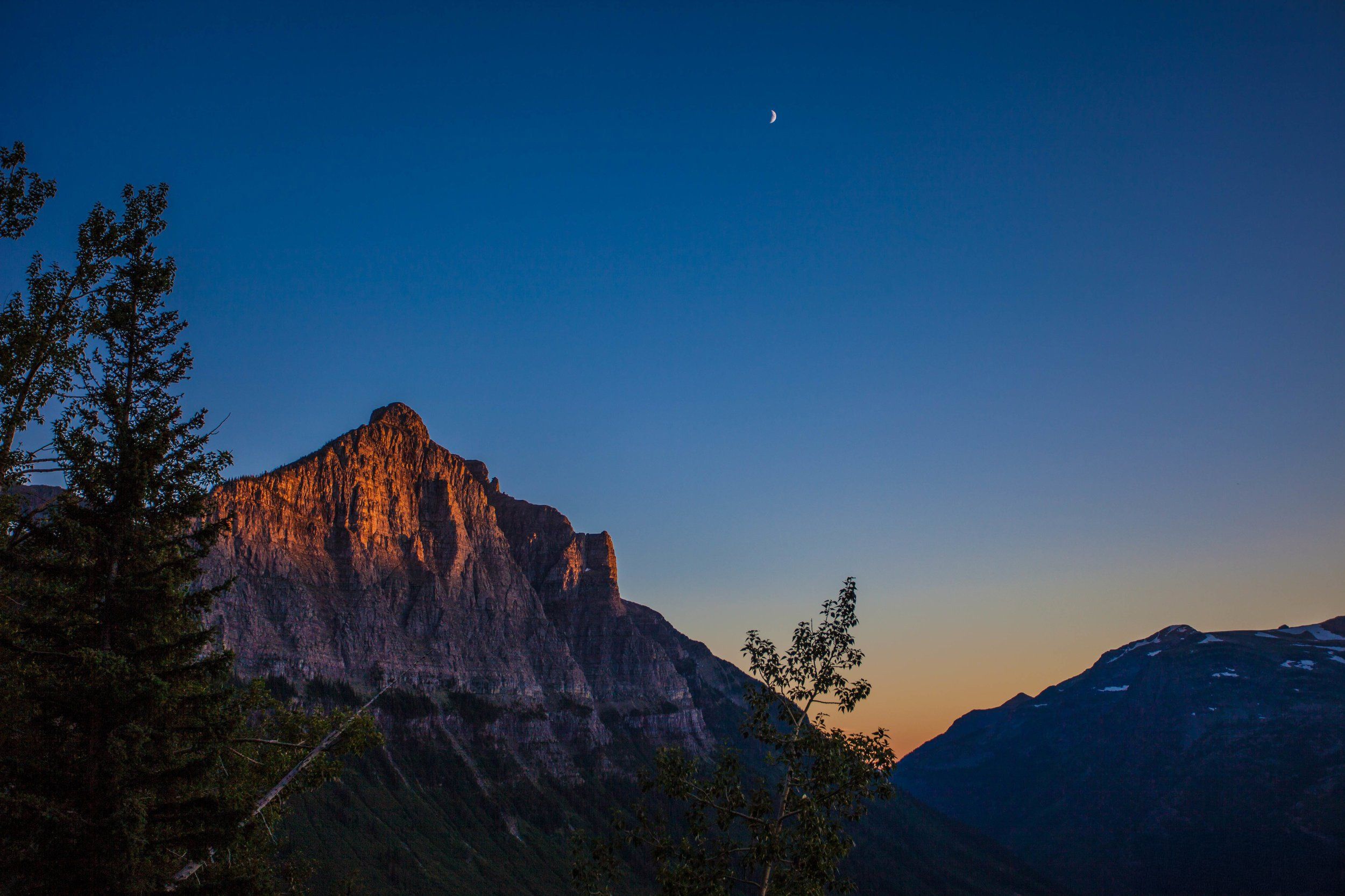 The last sunlight turning a rocky peak orange, dark pines below and a thin crescent moon in the deep blue sky.