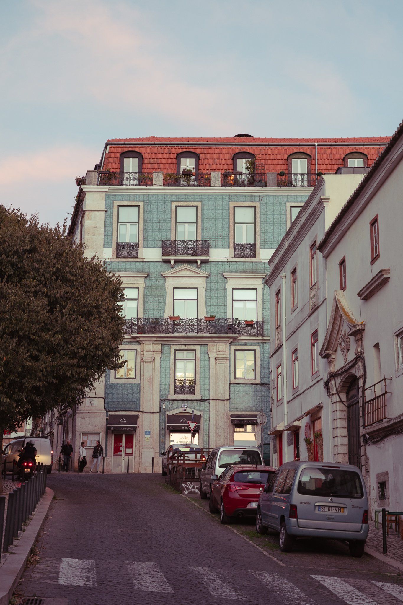 A blue-green tiled building with iron balconies and a red roof at the top of a sloping street, under a soft pink and blue sky.